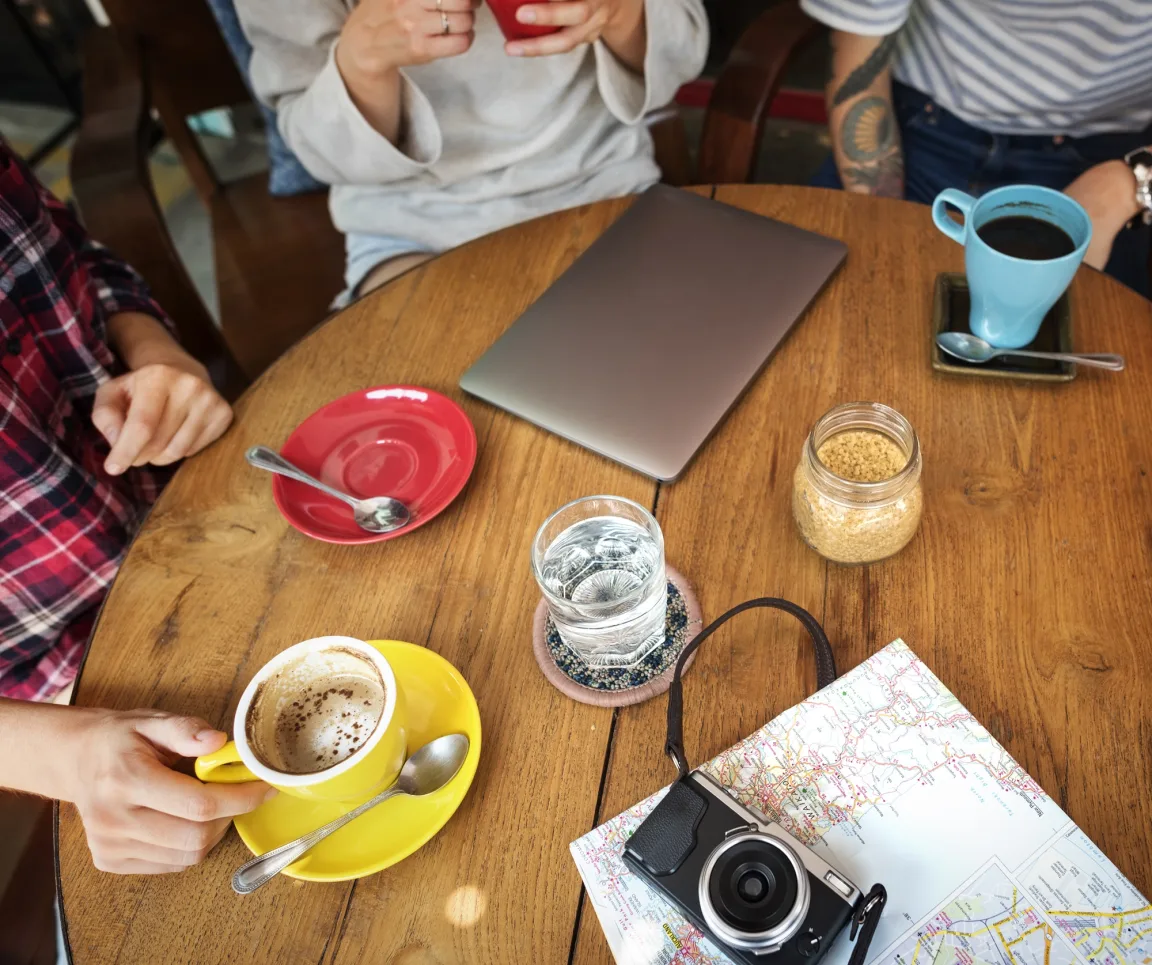 A group of friends planning a trip together at a bright table cluttered with maps, phones and laptops.